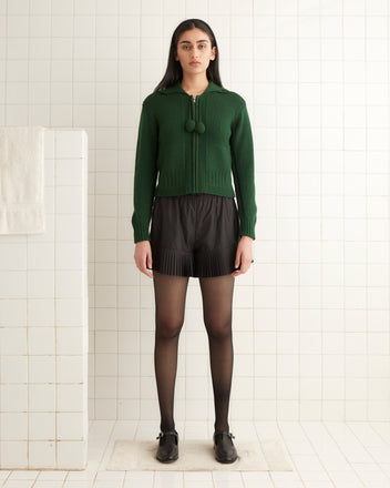 A woman in a green Marion Tassel Cardigan and black shorts stands in a tiled room, wearing black tights and loafers.