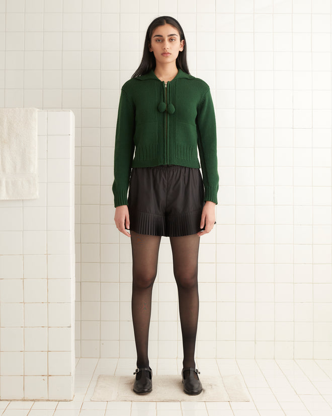 A woman in a green Marion Tassel Cardigan and black shorts stands in a tiled room, wearing black tights and loafers.