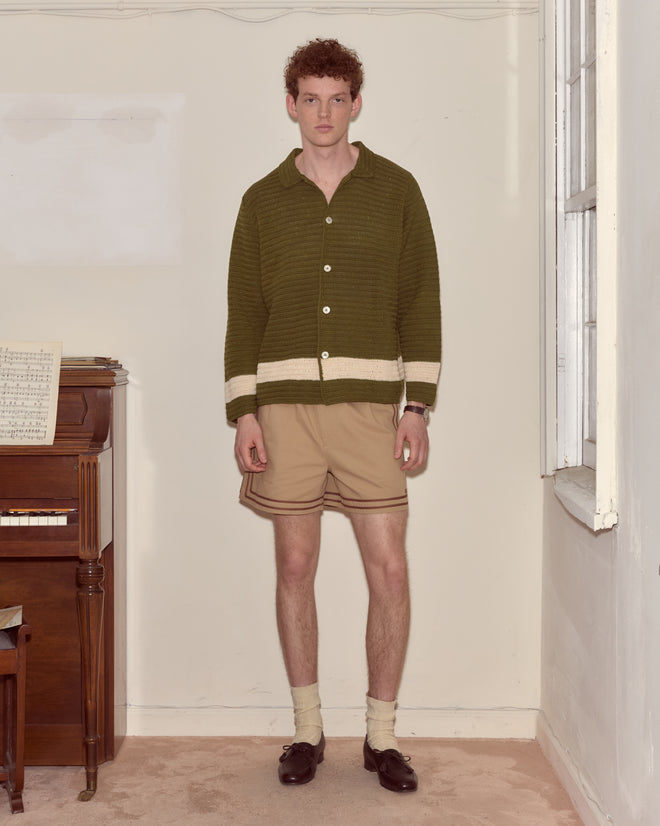 A young man in a green sweater and beige shorts stands indoors beside a piano and a window, his look complemented by the Marsh Crochet Long Sleeve Shirt draped over his shoulder.