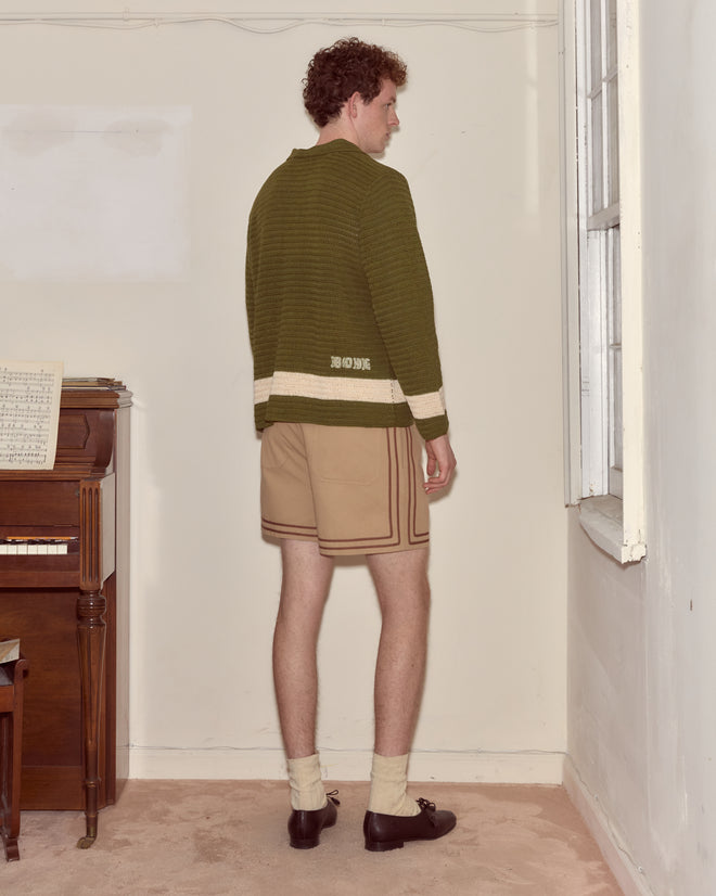 A man in a Marsh Crochet Long Sleeve Shirt and beige shorts stands near a window beside a piano with sheet music.