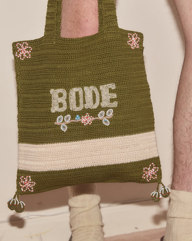 The Marsh Crochet Tote, featuring green cotton crochet and intricate floral beadwork with "BODE," is shown held by a person in cream socks.