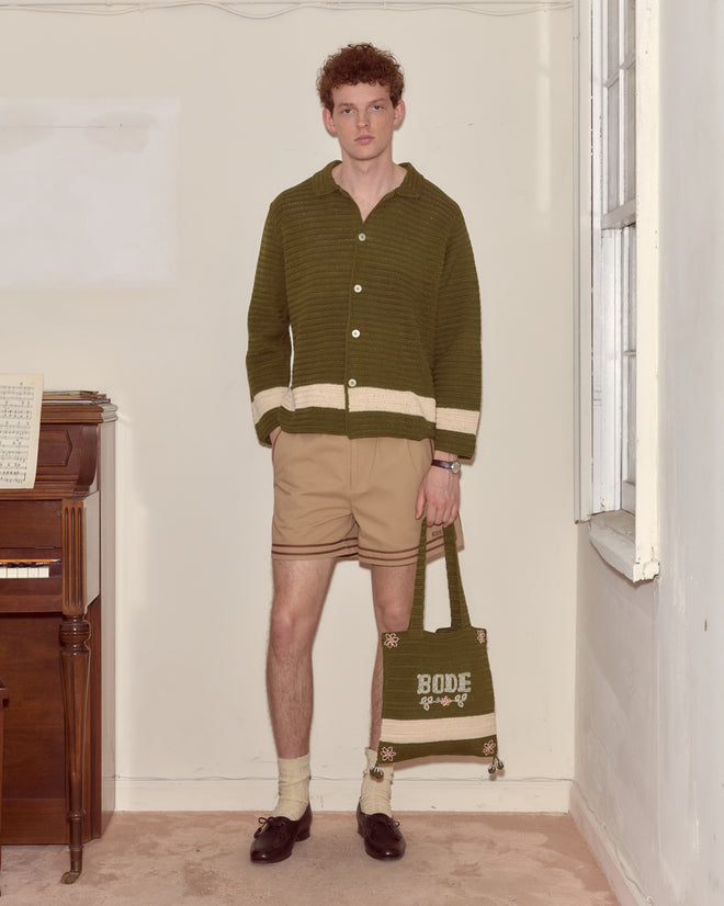 A man in a green sweater, tan shorts, and loafers holds the Marsh Crochet Tote by a piano.