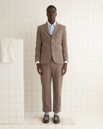 A person with long hair is wearing the Marston Check Suit Jacket paired with a white shirt made from 100% merino and brown loafers, standing against a light yellow wall.