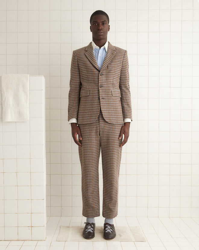 A person with long hair is wearing the Marston Check Suit Jacket paired with a white shirt made from 100% merino and brown loafers, standing against a light yellow wall.