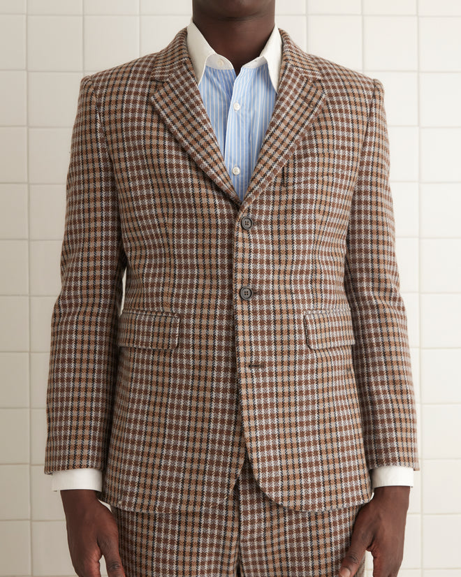 A person with long hair is wearing the Marston Check Suit Jacket paired with a white shirt made from 100% merino and brown loafers, standing against a light yellow wall.