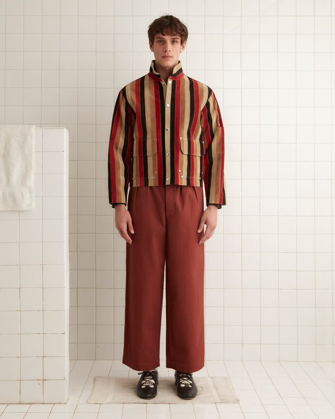 A person in the Matchstick Suede Jacket and rust-colored pants stands on a white tiled floor near a towel hanging on the wall.