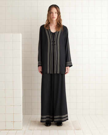 A woman wearing the Mathilde Jacket, which features black fabric with white striped details, stands on a white tiled floor.