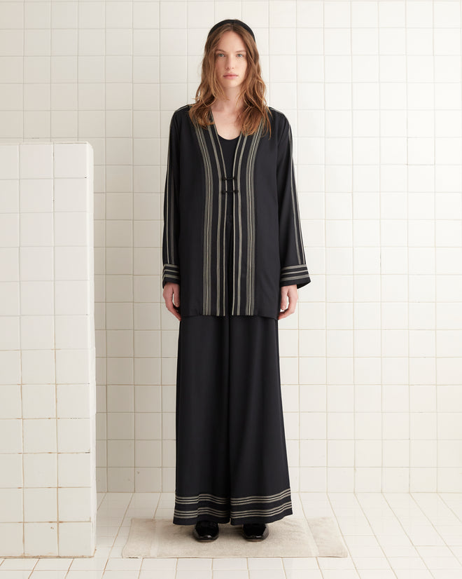 A woman wearing the Mathilde Jacket, which features black fabric with white striped details, stands on a white tiled floor.