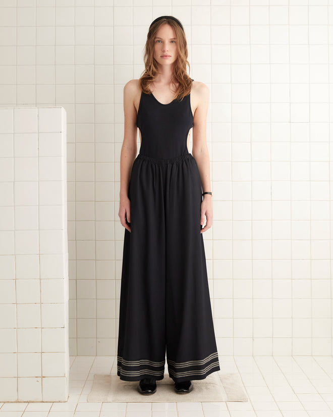 A woman wearing the Mathilde Trousers stands against a white tiled wall, her style reflecting the elegance of a 1940s peignoir.