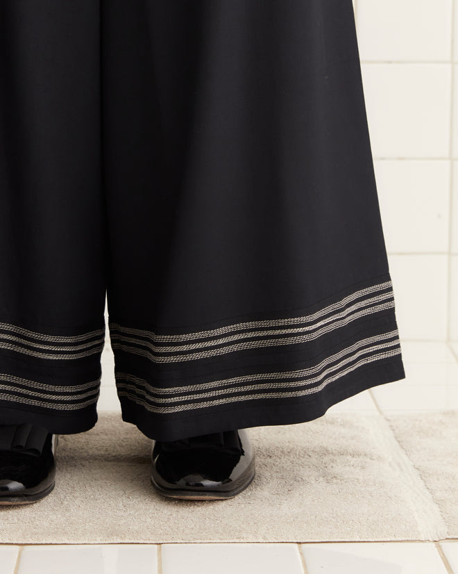 A woman wearing the Mathilde Trousers stands against a white tiled wall, her style reflecting the elegance of a 1940s peignoir.