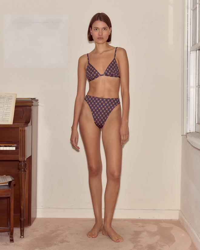 Woman wears McAllister Bikini Bottoms with a foulard print, standing indoors near a piano and window, facing the camera.