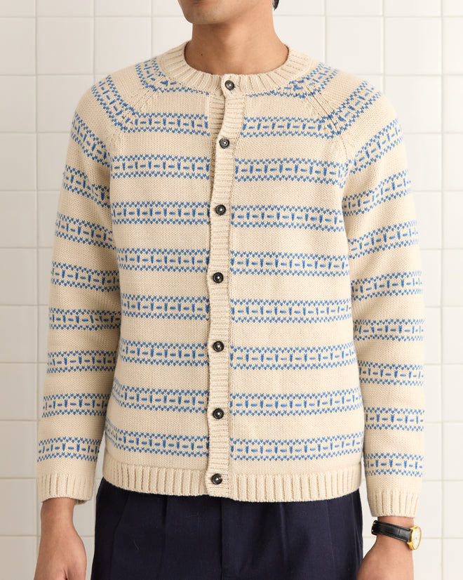 A person wearing the Meandering Stripe Cardigan - Cream, featuring a cream and blue geometric stripe pattern with brown buttons, stands against a tiled wall.