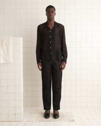 A person stands in a tiled room wearing the Meandering Lace Long Sleeve Shirt, a black sheer, button-up shirt crafted from 100% nylon, paired with black pants, looking directly at the camera.