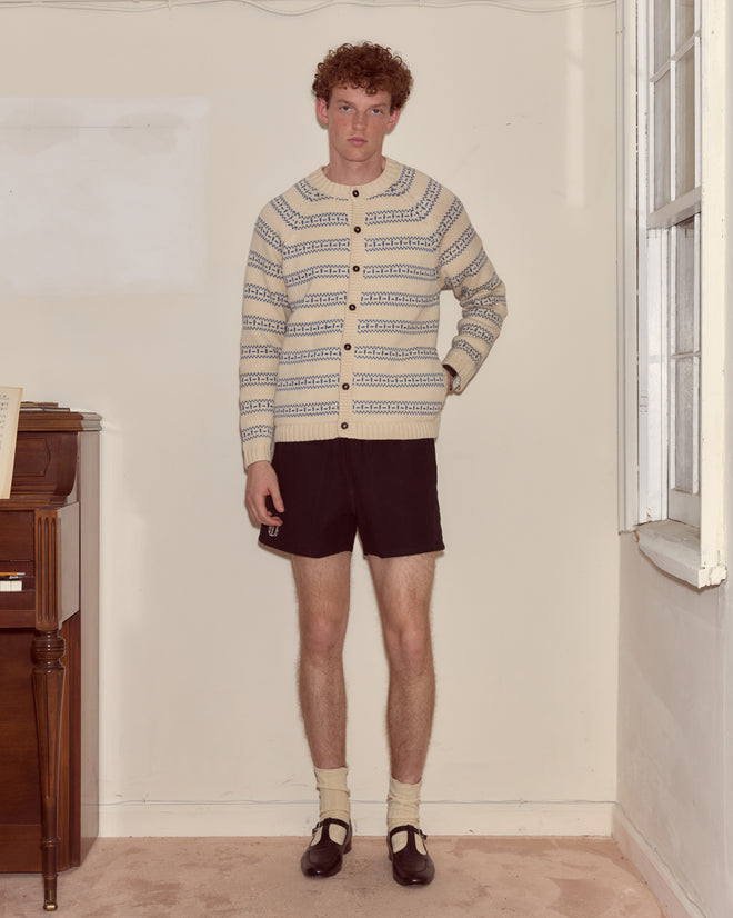 A young person wearing the Meandering Stripe Cardigan in cream, paired with black shorts, socks, and brown shoes, stands in a plain room by a window.