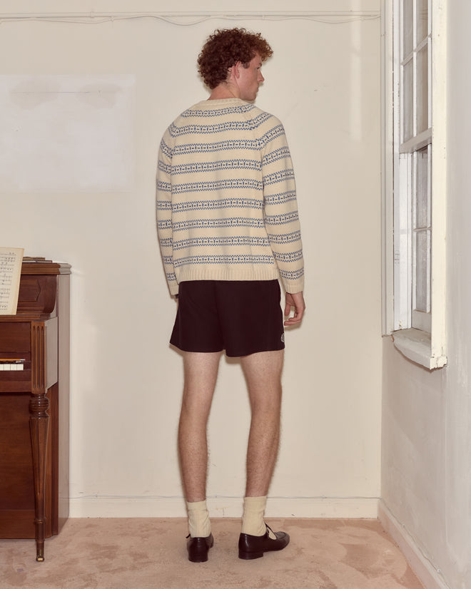 A person with curly hair, wearing the Meandering Stripe Cardigan - Cream and shorts, stands facing a wall in a sparsely furnished room.