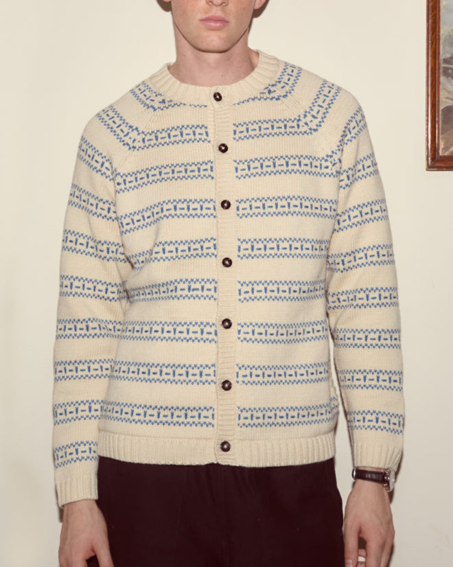 A person wearing the Meandering Stripe Cardigan - Cream with geometric blue stripes stands against a plain wall, their face partially visible.