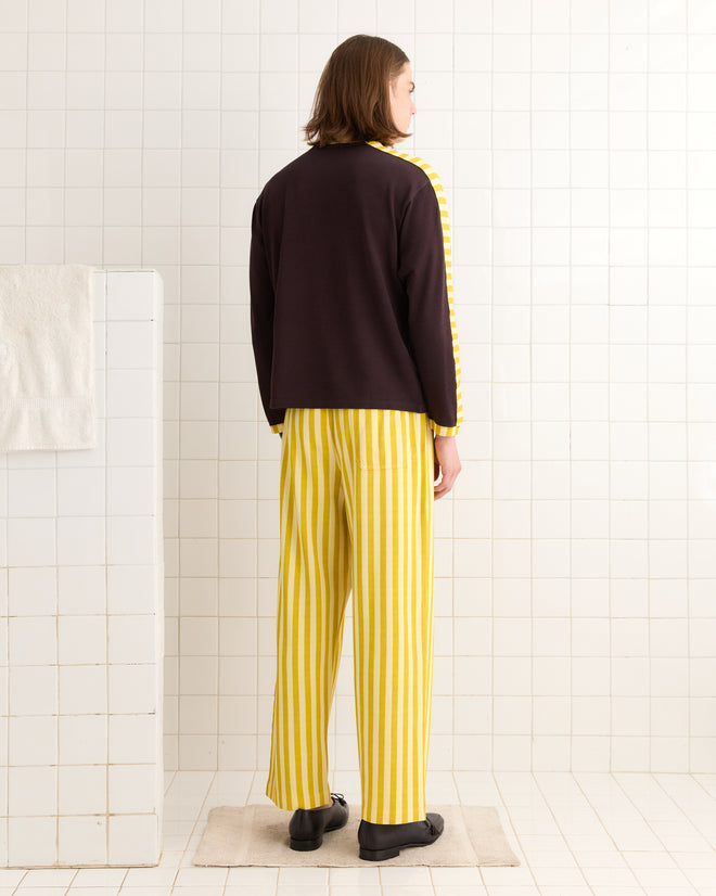 A person in a stylish outfit, featuring a heavyweight Medalist Appliqué Long Sleeve Tee and yellow striped pants, stands confidently on a rug in a white-tiled room.