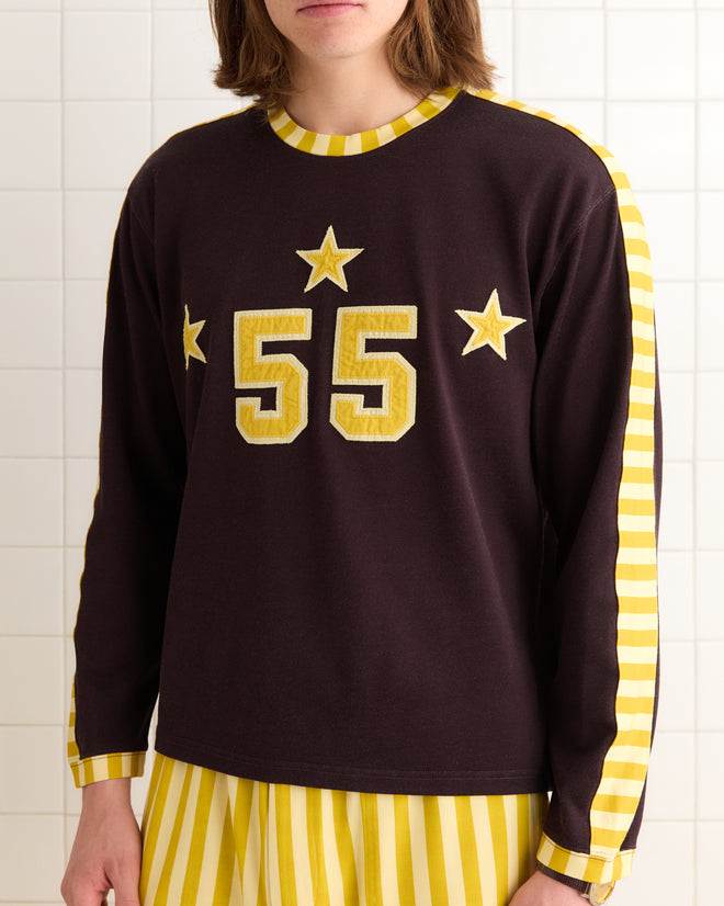 In front of a white tiled wall, someone is wearing the Medalist Appliqué Long Sleeve Tee, featuring "55" and stars on striped sleeves, crafted in a 1940s style from heavyweight fabric.