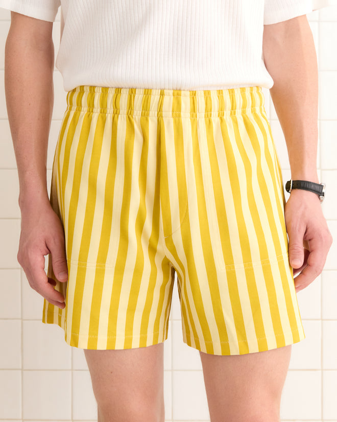 Wearing Medalist Shorts in yellow and white stripes paired with a classic white shirt, the person stands against a tiled backdrop, showcasing an effortless classic silhouette.