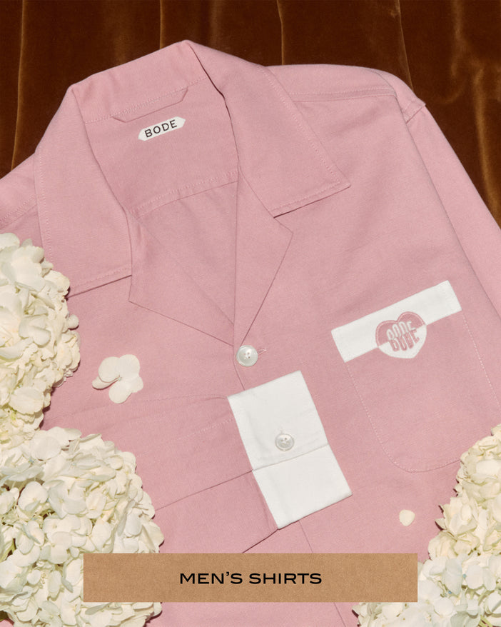 A pink and white button up shirt with a Bode heart logo on the front pocket. The shirt sits on brown velvet, surrounded by white flowers.
