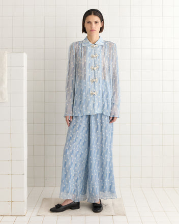 A person in a light blue patterned Meridian Blouse reminiscent of Art Deco style stands against a tiled wall in black shoes. Their short hair complements the vintage vibe, blending modern flair with 1930s geometric elements.