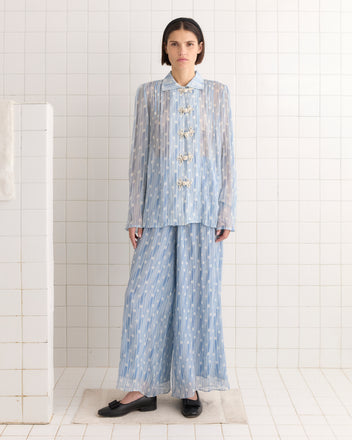 Dressed in the Meridian Blouse, a light blue patterned outfit with large buttons reflecting 1930s Art Deco style, a person stands in a tiled room. The ensemble is completed with black shoes, adding an elegant geometric flair reminiscent of the era.