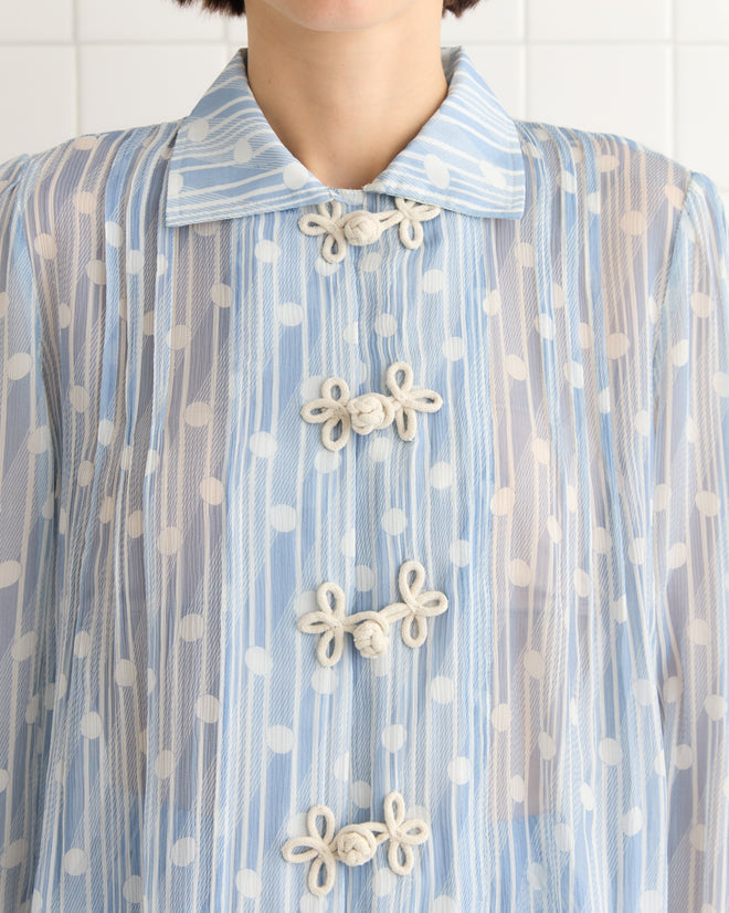 A person wearing the Meridian Blouse, a sheer light blue silk top adorned with polka dots and intricate knotted buttons.