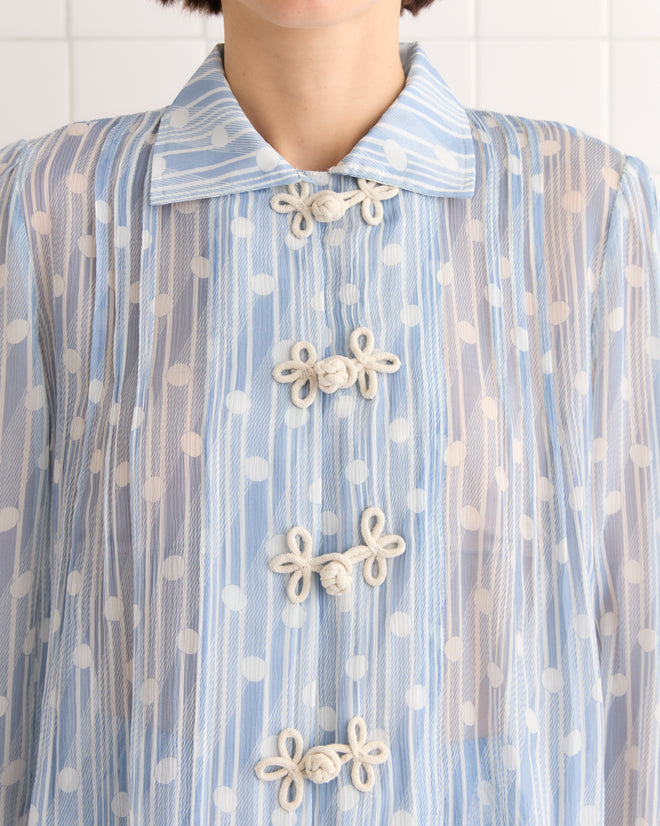 Close-up of a person wearing the Meridian Blouse, a sheer blue 100% silk top with white polka dots and decorative knot buttons, inspired by 1930s Art Deco geometric style.