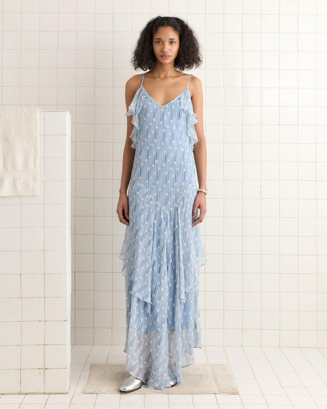 In a tiled room, someone stands wearing a Meridian Gown, featuring blue patterns and shoulder straps, with a towel nearby.