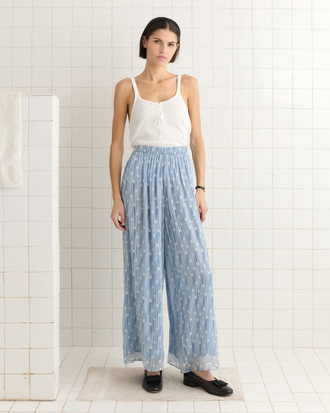 A person wearing a white tank top and Meridian Pants, featuring a blue pattern and wide-leg silhouette, stands in a white-tiled room.