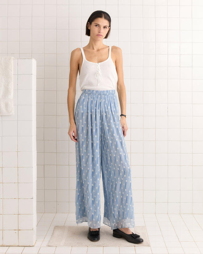 A woman in a white tank top and Meridian Pants stands gracefully in a tiled room.