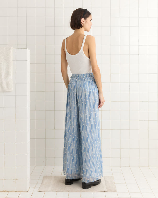 A woman in a white tank top and Meridian Pants, featuring striking blue silk with an Art Deco pattern, stands in a tiled room, facing away.