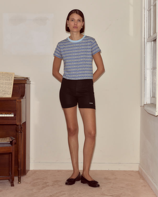 Indoors by a piano, a woman wears black Meringue Shorts, a blue knit top, and black flats.