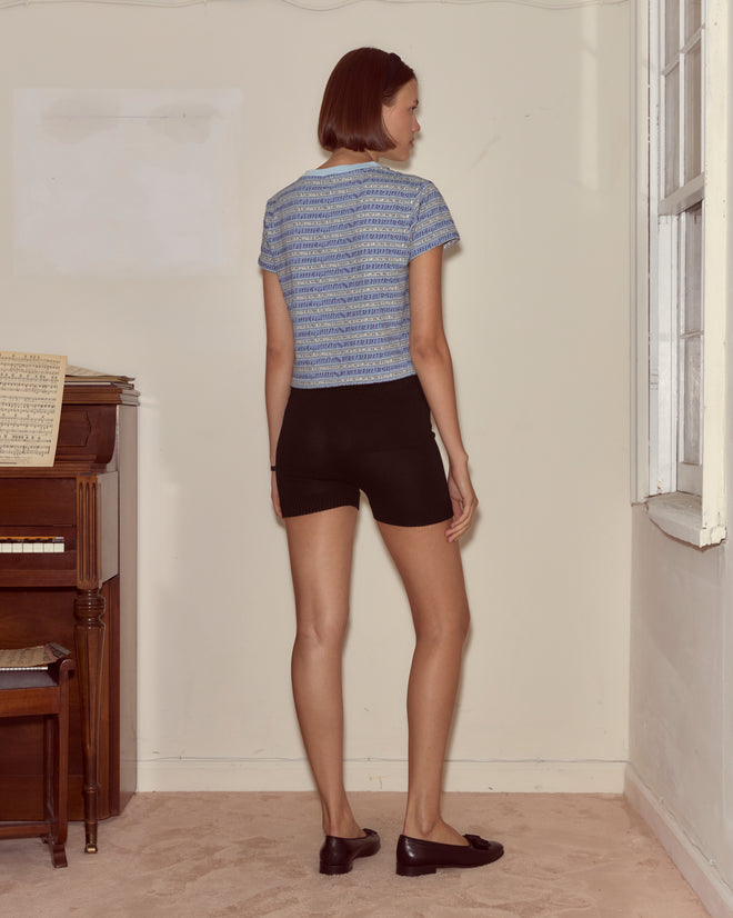A woman stands indoors by a piano, facing away, wearing a blue shirt, black Meringue Shorts and black shoes.