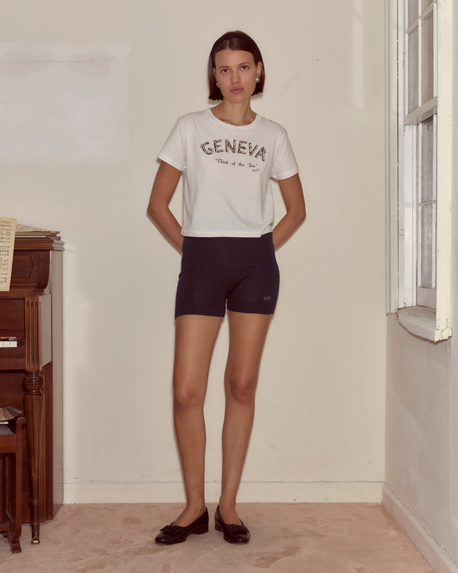 Woman stands indoors wearing a "GENEVA" t-shirt, Meringue Shorts - Blue, and black flats beside a piano and window.