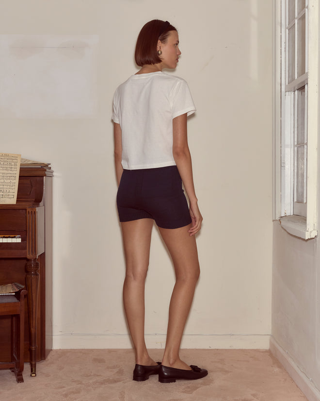 A woman stands indoors by a window and a piano, wearing a white shirt and Meringue Shorts in blue, facing away.