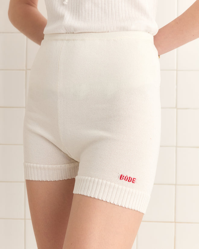 A person stands against a tiled wall, wearing Meringue Shorts - Ivory, crafted from soft cotton yarn. "CODE" is boldly emblazoned in red on the hem, with a retro-inspired design nodding to the 1960s-1970s style.