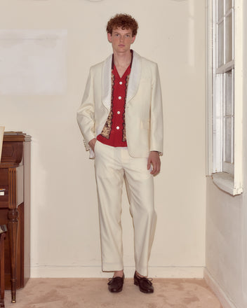 A man wearing the Meringue Shawl Collar Tuxedo Jacket in ivory with a red shirt stands indoors beside a piano and window, looking at the camera.