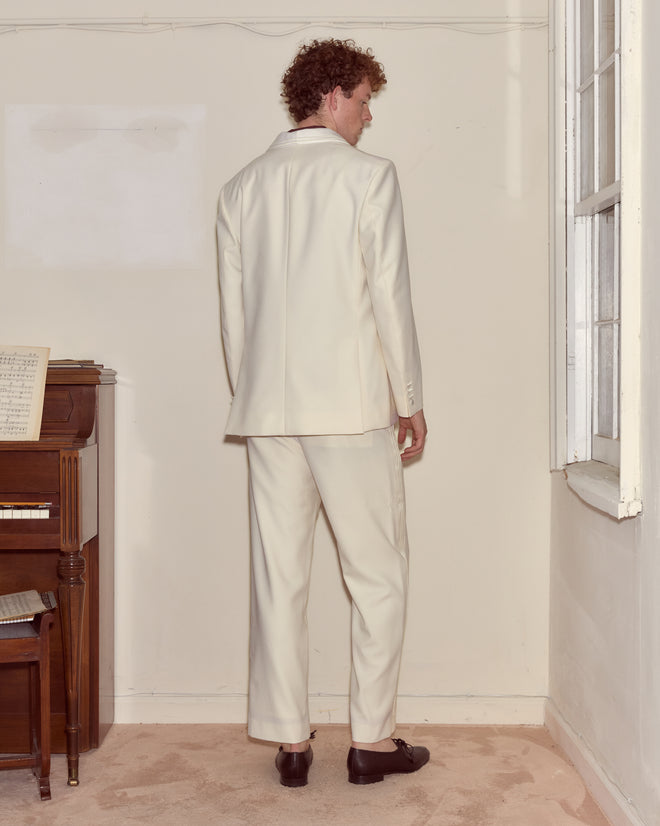A man wearing a Meringue Shawl Collar Tuxedo Jacket stands facing the corner of a beige room near a window and a piano.