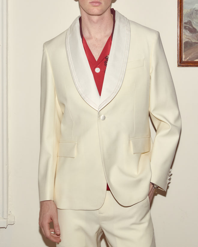 A person stands indoors, partially out of frame, wearing the Meringue Shawl Collar Tuxedo Jacket in ivory wool over a red shirt.