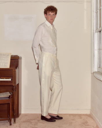 A man in white formal attire stands indoors by a window near a piano, looking at the camera, dressed in Meringue Smoking Trousers—ivory wool trousers with a satin side stripe for classic elegance.
