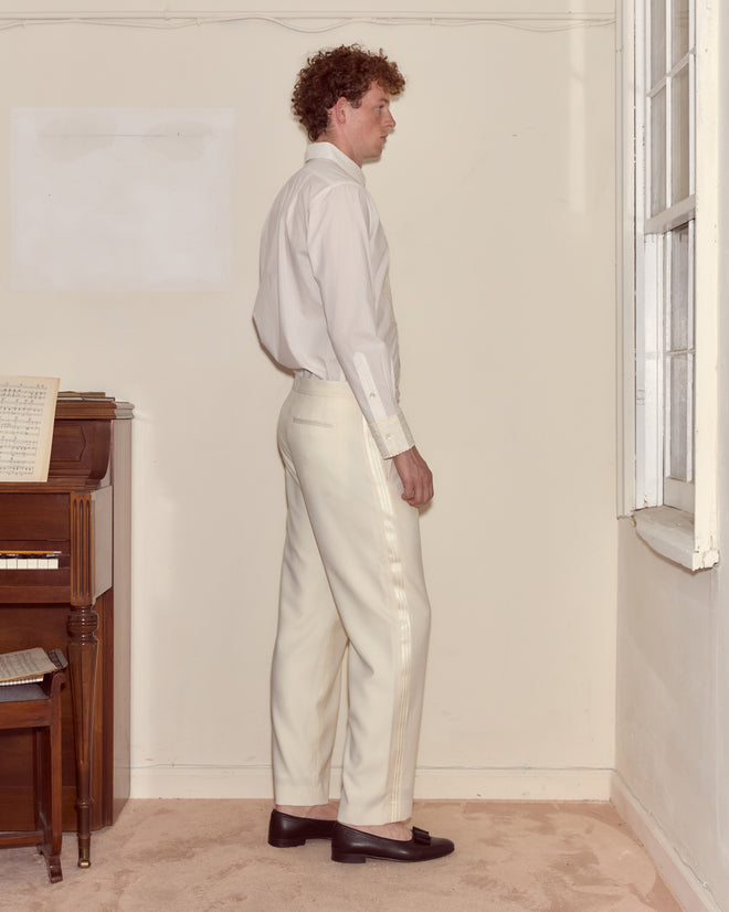 A man wearing Meringue Smoking Trousers and a white shirt stands sideways by a window in a sparsely furnished room.