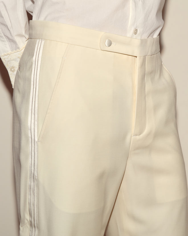 Wearing Meringue Smoking Trousers in ivory wool with a satin side stripe, paired with a crisp white buttoned shirt.