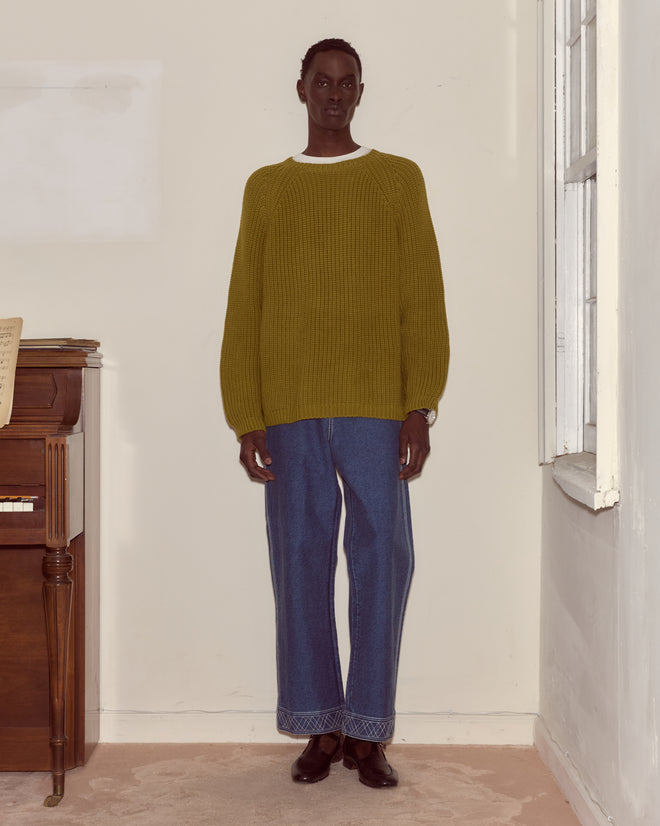 A person wears the Merton Crewneck in olive green with blue jeans, standing indoors near a piano and a window, facing the camera.