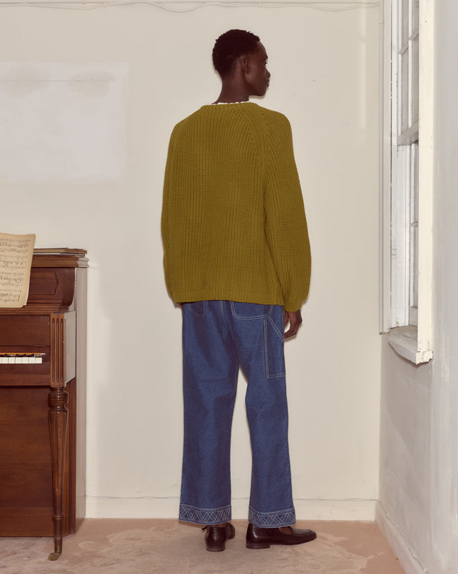 Wearing the Merton Crewneck in green, made from a cozy cotton merino blend, and blue jeans, a person stands facing a wall by a piano and window.