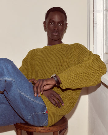 Wearing the Merton Crewneck in mustard shaker stitch and blue jeans, a person sits on a stool by the window, looking confidently at the camera.