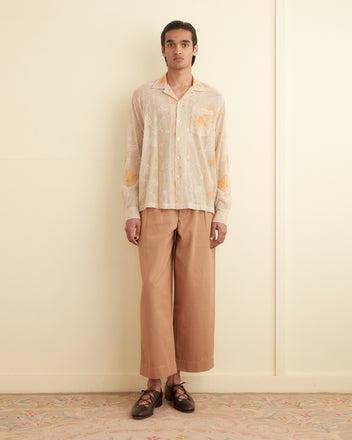 A person stands against a plain wall, wearing the Mesh Lilium Long Sleeve Shirt, featuring hand-guided embroidery, and beige wide-leg pants. The floor is adorned with a light-colored rug.