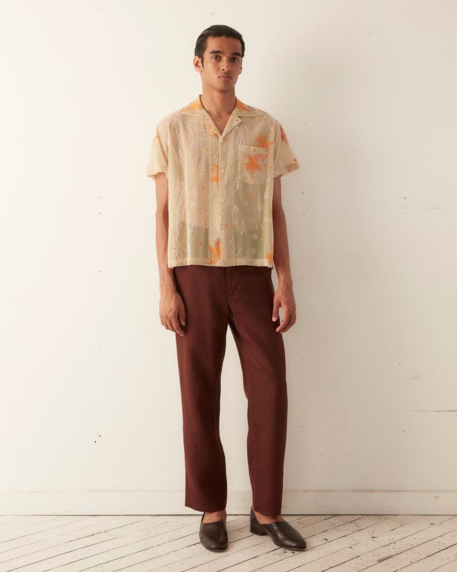 A person stands against a plain white wall, wearing the Mesh Lilium Short Sleeve Shirt with hand-guided embroidery and brown pants, paired with brown shoes.