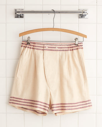 Metric Redbud Boxers, featuring a beige base with red and blue patterned trim, hung on a wooden hanger against a white tiled wall, exude a one-of-a-kind charm. The subtle hand-mending gives them an added touch of uniqueness.