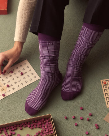 A person wearing Micro Check Socks - Purple, crafted in Italy, sits on the floor, enthusiastically playing bingo with markers and a card.